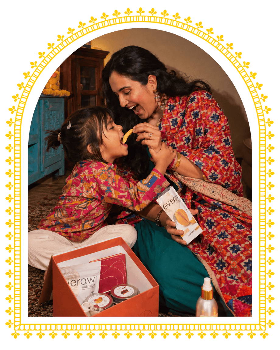 A mother laughs as she feeds her little daughter an Everaw mango snack beside an open Everaw Diwali gift box, framed in a golden festive arch